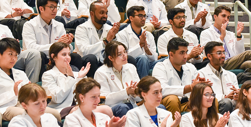 Medical students wearing white coats sitting in an auditorium and clapping.