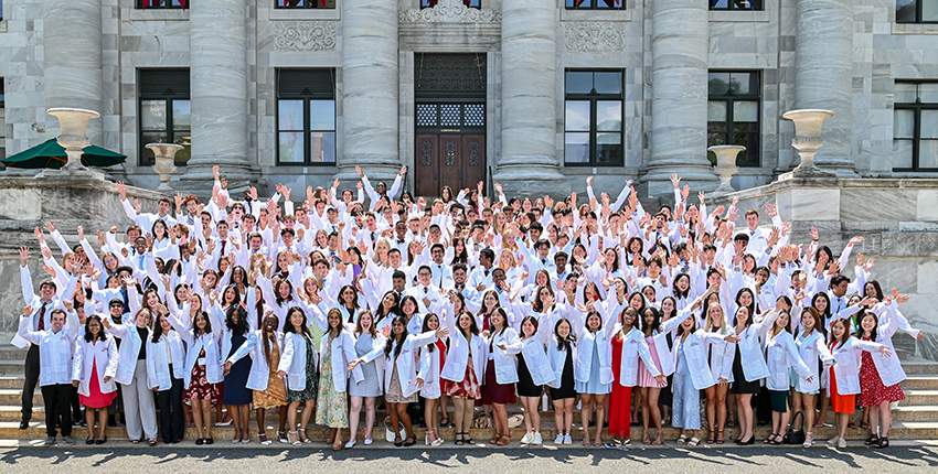 Over 100 students wearing white coats waving their arms in excitement on the steps leading to a marble building.
