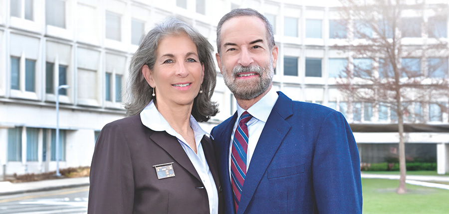 Woman and man smiling in front of a building with a tree in the background.