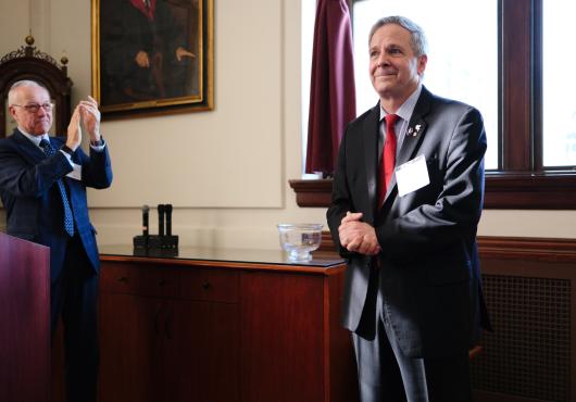 Man standing with his hands clasped in front of him with a humble smile and a man clapping in the background.