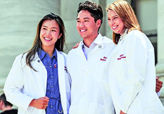 Three medical students wearing white coats with "Harvard Medical School" embroidered on them, with the marble pillars of Gordon Hall in the background.