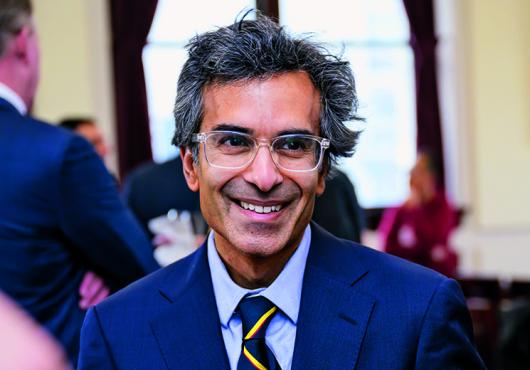 Man wearing a suit and glasses smiling with a crowd behind him.