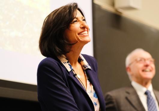 Woman giving a presentation in front of a large screen with a man smiling in the background.
