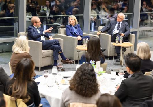 Photo of 3 people sitting in chairs speaking in front of tables 