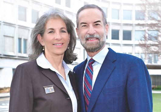 Woman and man smiling in front of a building with a tree in the background.