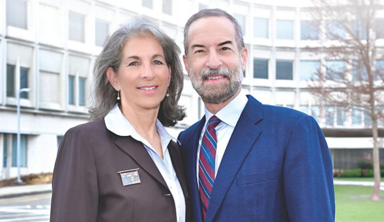 Woman and man smiling in front of a building with a tree in the background.